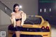 A woman posing in front of a yellow sports car.