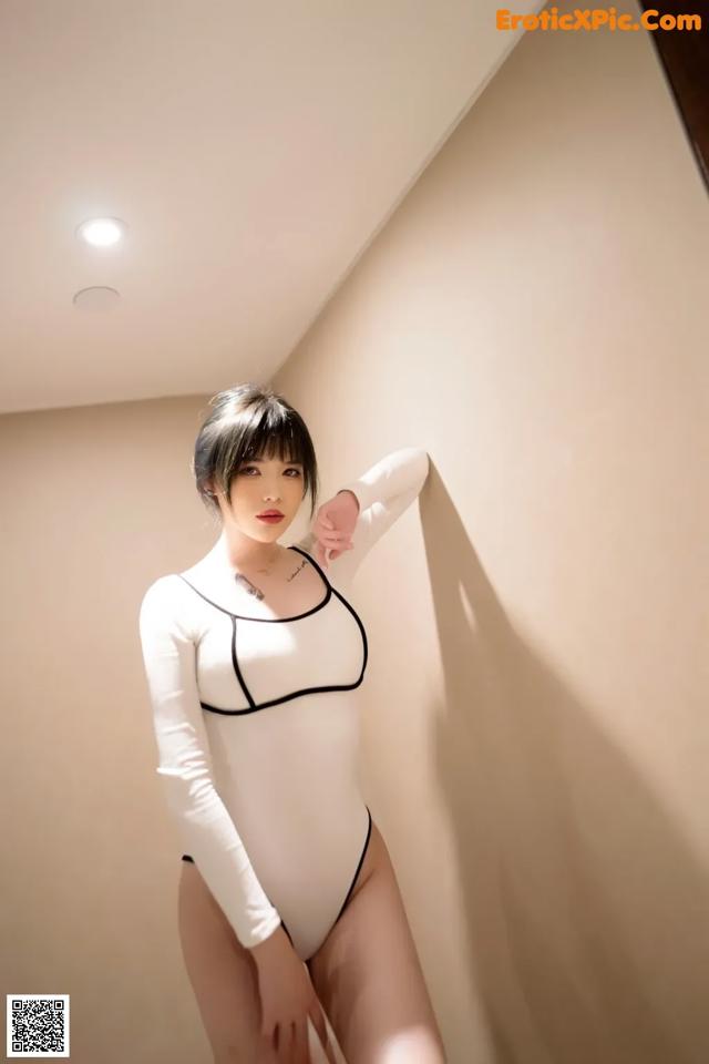 A woman in a white bodysuit leaning against a wall.