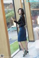 A woman in a black shirt and blue skirt standing by a window.